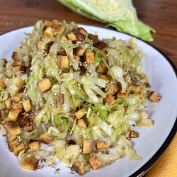 Auf dem Teller liegt eine Kombination aus knackigem Spitzkohl, goldbraunem Tofu und geröstetem Sesam. Im Hintergrund betont ein angeschnittener Spitzkohl die Frische der Zutaten. Ein leichtes und gesundes Gericht mit appetitlicher Optik.
