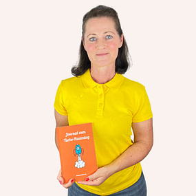 Sonja Fuchs alias Fuchsmunter hält das orangefarbene „Journal zum Turbo-Fastentag“ in den Händen. Sie trägt ein gelbes Shirt und steht vor hellem Hintergrund.